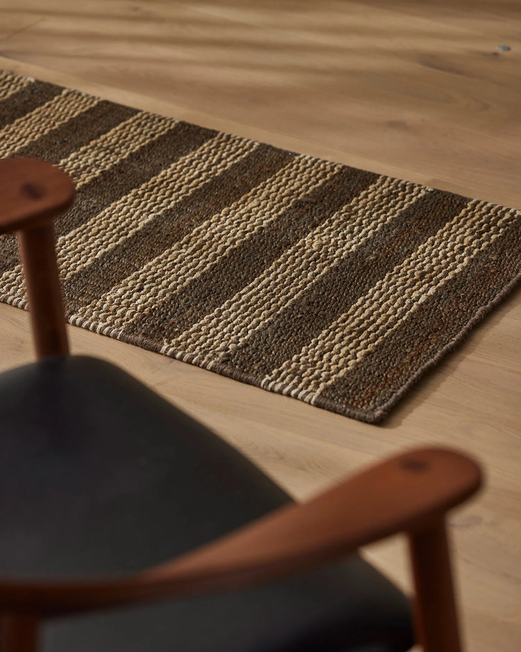 Neutral striped jute doormat in natural and charcoal tones, layered on light timber flooring beside mid-century wooden chair with dark upholstery